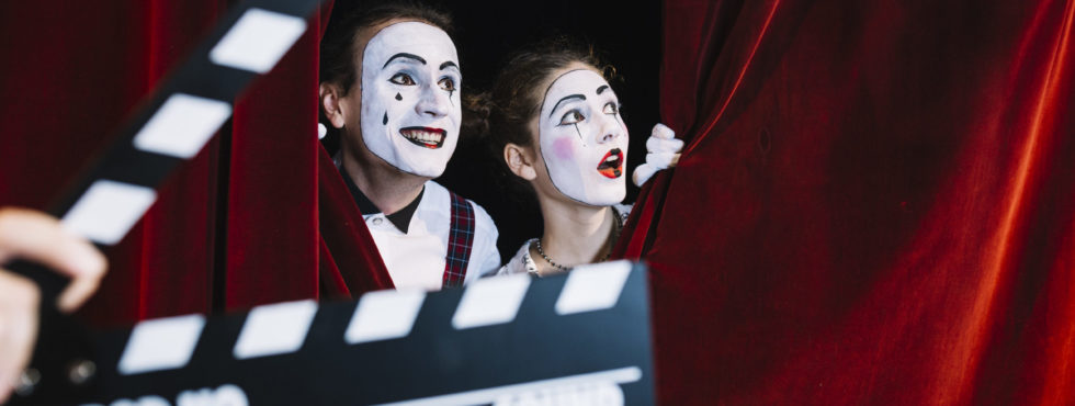 Clapperboard In Front Of Excited Mime Couple Peeking Behind The Red Curtain