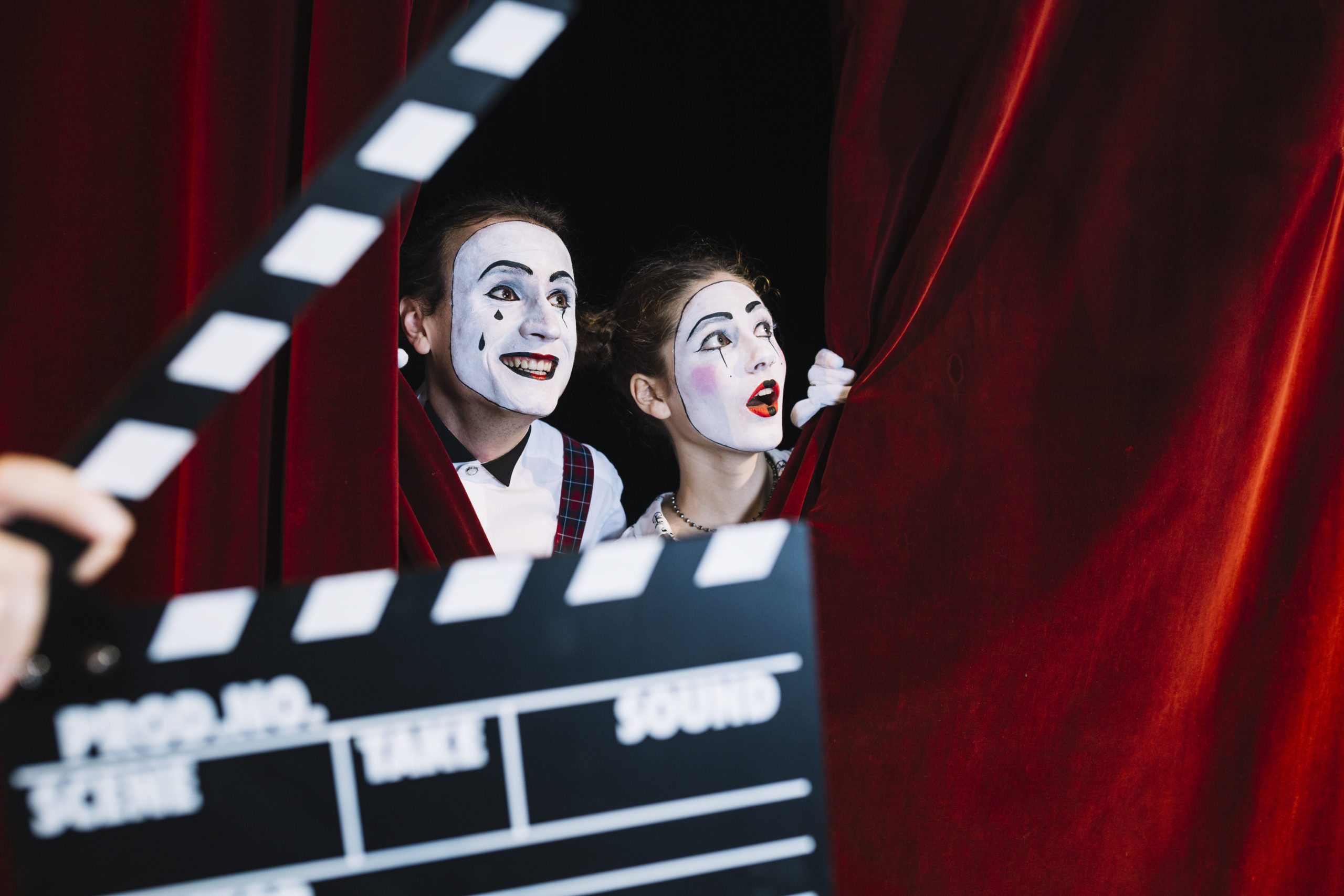 Clapperboard In Front Of Excited Mime Couple Peeking Behind The Red Curtain